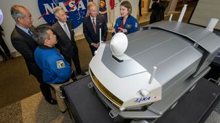 three men in suits and two astronauts wearing blue flight suits stand beside a model of a moon rover